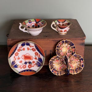 Può includere: Una collezione di tazze da tè, piattini e un piatto da portata in porcellana antica. I pezzi presentano intricati motivi floreali in arancione, blu e oro su sfondo bianco. Le tazze hanno piccole maniglie e sono su una superficie di legno.