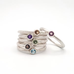 Könnte beinhalten: Sechs Silberringe mit bunten Edelsteinen übereinander gestapelt. Die Ringe sind übereinander gestapelt, wobei die Edelsteine nach außen zeigen. Die Edelsteine sind Amethyst, Grün, Braun, Blau, Gelb und Rosa.