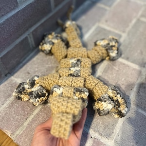 May include: A handmade crochet lizard toy in tan, gray, and cream colors. The lizard is lying on a brick surface. The toy has a segmented body and four limbs, with a tail extending from the body.