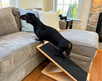 Pode incluir: Um cão dachshund preto usando uma rampa de madeira para animais de estimação para subir em um sofá cinza. A rampa tem uma superfície preta antiderrapante.