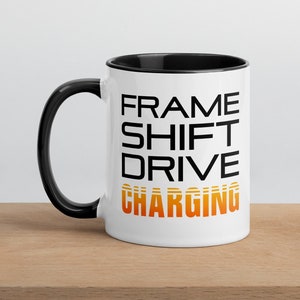 Puede incluir: Taza de cerámica blanca con borde y asa negros. La taza tiene texto negro que dice "FRAME SHIFT DRIVE CHARGING" con la palabra "CHARGING" en naranja y amarillo.