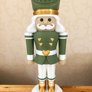 Könnte beinhalten: Eine Nussknacker-Figur in einer matten olivgrünen Uniform mit goldenen herzförmigen Akzenten auf Hut, Brust und Stiefeln. Die Figur hat einen weißen Schnurrbart, weißes Haar, eine weiße Hose und steht auf einem weißen herzförmigen Sockel.