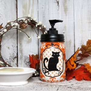 May include: Orange and white Halloween themed soap dispenser with a black cat design. The dispenser is made from a repurposed glass jar with a black pump top.