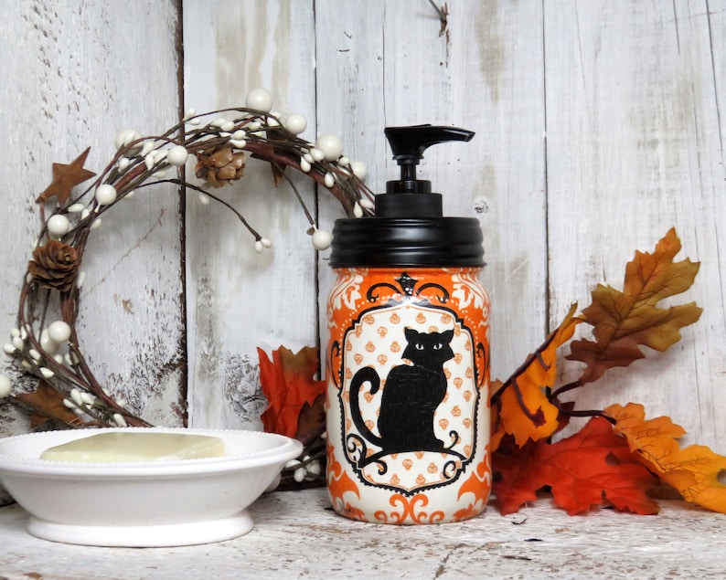 May include: Orange and white Halloween themed soap dispenser with a black cat design. The dispenser is made from a repurposed glass jar with a black pump top.