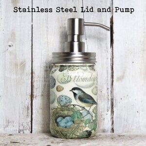 May include: A glass soap dispenser with a stainless steel lid and pump. The dispenser is decorated with a bird and nest design. The text "Hommede" is visible on the dispenser.