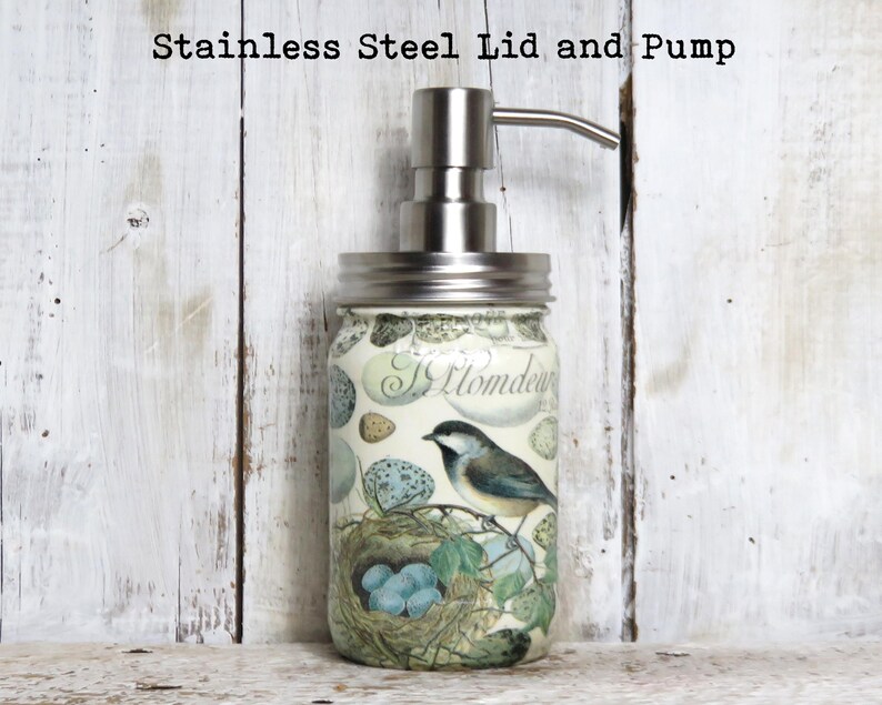 May include: A glass soap dispenser with a stainless steel lid and pump. The dispenser is decorated with a bird and nest design. The text "Hommede" is visible on the dispenser.
