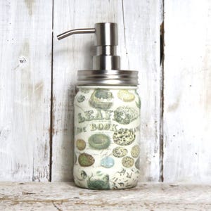 May include: A white glass soap dispenser with a stainless steel pump and a vintage-inspired design featuring a botanical illustration. The dispenser is labeled "Leaves from the Book of Nature".
