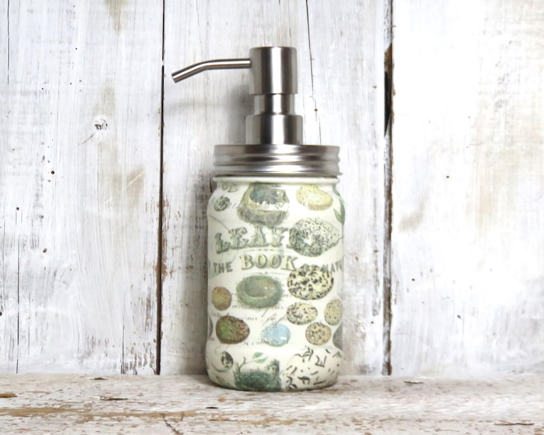 May include: A white glass soap dispenser with a stainless steel pump and a vintage-inspired design featuring a botanical illustration. The dispenser is labeled "Leaves from the Book of Nature".