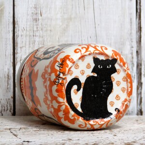 May include: A white ceramic jar with an orange and black Halloween design. The jar features a black cat silhouette with a white background and a repeating pattern of orange skulls on a white background. The jar has a silver lid.