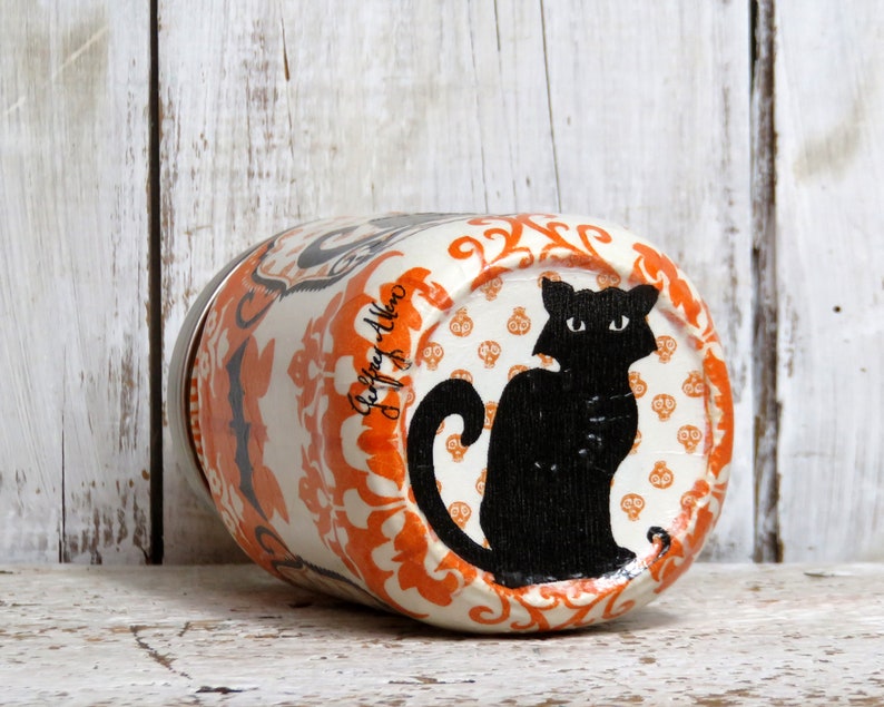 May include: A white ceramic jar with an orange and black Halloween design. The jar features a black cat silhouette with a white background and a repeating pattern of orange skulls on a white background. The jar has a silver lid.