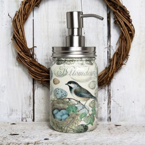 May include: A glass soap dispenser with a stainless steel pump and lid. The dispenser is decorated with a vintage-style illustration of a bird's nest with eggs and a bird perched on a branch. The text "J. Plomdeur" is visible on the front of the dispenser.