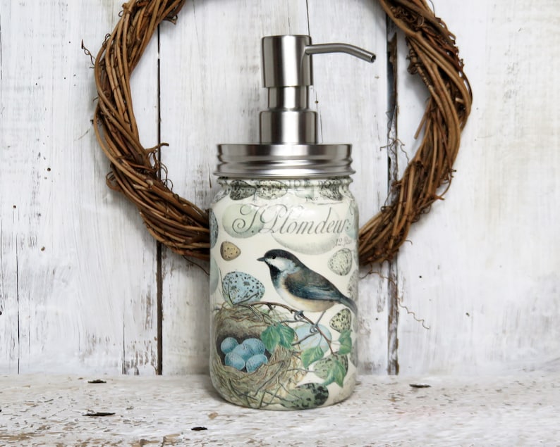 May include: A glass soap dispenser with a stainless steel pump and lid. The dispenser is decorated with a vintage-style illustration of a bird's nest with eggs and a bird perched on a branch. The text "J. Plomdeur" is visible on the front of the dispenser.