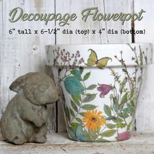 May include: A white ceramic flowerpot with a decoupage design of pressed flowers, leaves, and a butterfly. The pot is 6 inches tall, 6 1/2 inches in diameter at the top, and 4 inches in diameter at the bottom.  The pot is sitting on a white wooden surface next to a small gray concrete rabbit figurine.