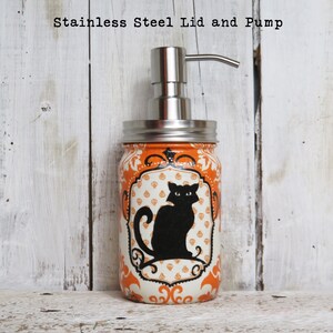May include: Orange and white mason jar soap dispenser with a black cat design and a stainless steel lid and pump. "Stainless Steel Lid and Pump" is written above the jar.