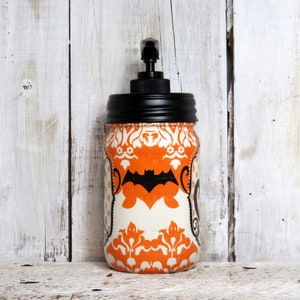May include: Orange and white Halloween themed soap dispenser with a black pump and a black bat design. The dispenser is in a glass jar with a black lid.