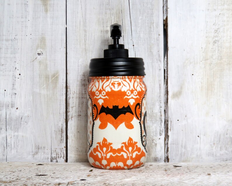 May include: Orange and white Halloween themed soap dispenser with a black pump and a black bat design. The dispenser is in a glass jar with a black lid.
