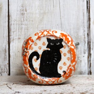 May include: A white ceramic round ornament with an orange border and a black cat silhouette on a white background with orange skull and crossbones patterns. The ornament has the text "Jeff & Allen" written on the side.