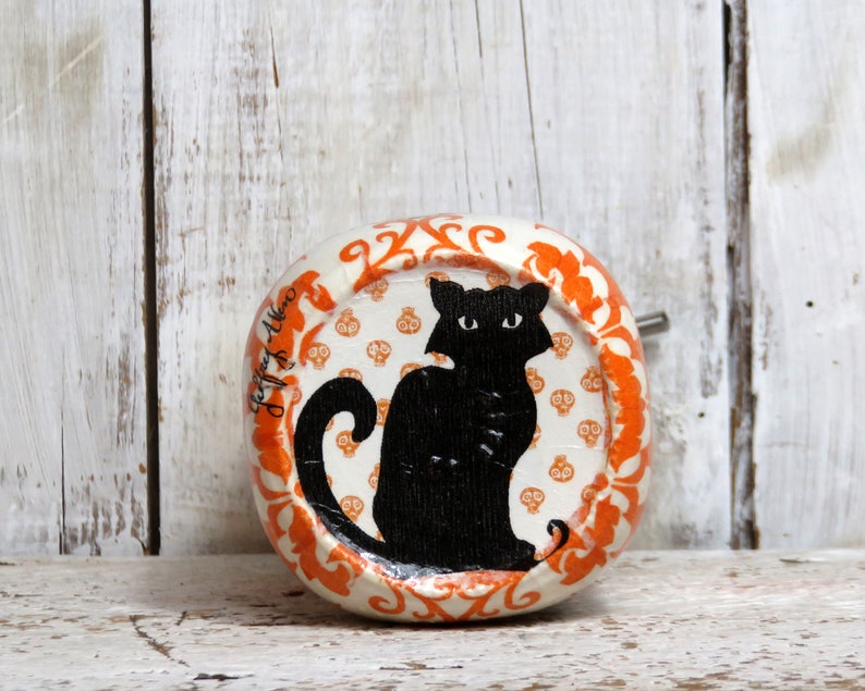 May include: A white ceramic round ornament with an orange border and a black cat silhouette on a white background with orange skull and crossbones patterns. The ornament has the text "Jeff & Allen" written on the side.
