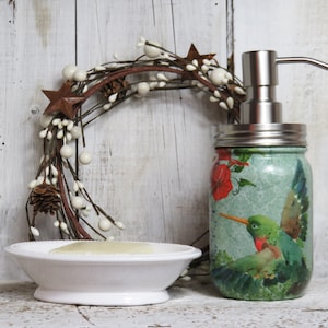 May include: A green glass jar with a stainless steel pump top and a hummingbird design. The jar is filled with liquid soap. A white ceramic soap dish is next to the jar.