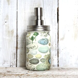 May include: A glass soap dispenser with a stainless steel pump and a white background with a pattern of blue and green eggs and the word "NATURE".