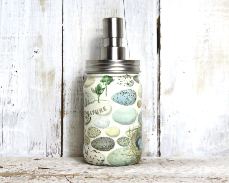 May include: A glass soap dispenser with a stainless steel pump and a white background with a pattern of blue and green eggs and the word "NATURE".