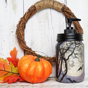 May include: A glass jar with a black lid and a pump dispenser. The jar is decorated with a spooky forest scene with a full moon and bats. The jar is sitting on a white wooden surface with a fall-colored leaf arrangement and a small orange pumpkin.