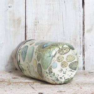 May include: A white glass jar with a silver lid. The jar is decorated with a pattern of green, blue, and brown stones. The jar is sitting on a white wooden surface.