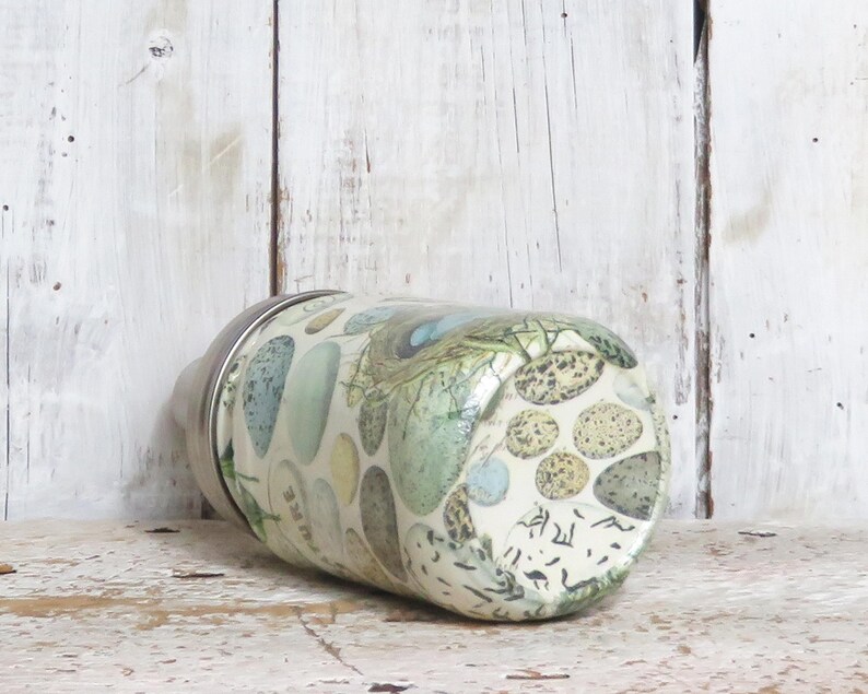 May include: A white glass jar with a silver lid. The jar is decorated with a pattern of green, blue, and brown stones. The jar is sitting on a white wooden surface.