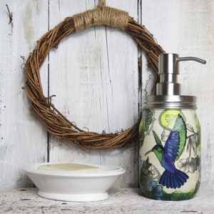 May include: A white ceramic soap dish with a bar of soap inside, a decorative glass soap dispenser with a hummingbird design, and a brown twig wreath hanging on a white wooden wall.