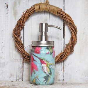 May include: A decorative soap dispenser made from a mason jar. The jar is wrapped in a colorful floral design featuring hummingbirds, pink flowers, and green leaves on a light blue background. The dispenser has a silver pump and lid. A brown wreath is behind the jar.