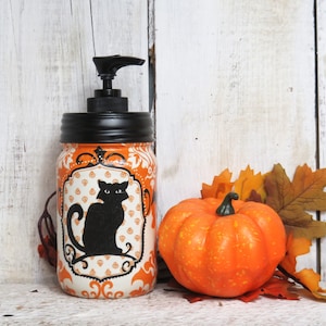 May include: Orange and white Halloween themed soap dispenser with a black cat silhouette. The dispenser is made from a glass jar with a black lid and a black pump. The dispenser is sitting on a white wooden surface with a small orange pumpkin and fall leaves.