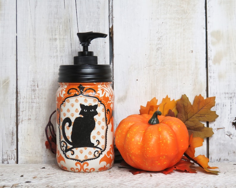 May include: Orange and white Halloween themed soap dispenser with a black cat silhouette. The dispenser is made from a glass jar with a black lid and a black pump. The dispenser is sitting on a white wooden surface with a small orange pumpkin and fall leaves.