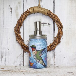 May include: A blue glass soap dispenser with a stainless steel pump and a hummingbird design. The dispenser is sitting on a white wooden surface.