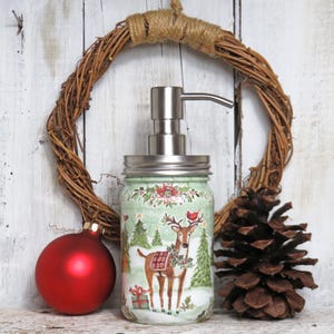 May include: A green glass soap dispenser with a silver pump and a Christmas-themed design featuring a deer, a bird, and a wreath. The dispenser is decorated with red, green, and white accents.