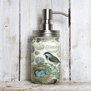 May include: A white ceramic soap dispenser with a silver pump and a floral design featuring a bird and nest. The design includes the text "Il Giardino" and a bird with blue and black feathers.