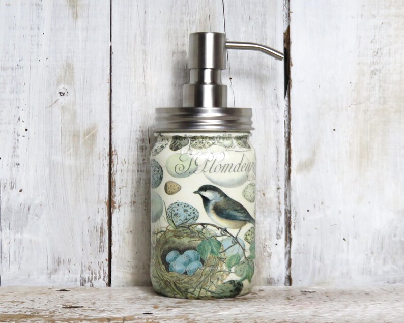 May include: A white ceramic soap dispenser with a silver pump and a floral design featuring a bird and nest. The design includes the text "Il Giardino" and a bird with blue and black feathers.