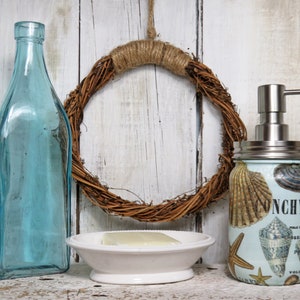 May include: A blue glass bottle, a white ceramic soap dish with a bar of soap, and a glass jar with a silver pump dispenser decorated with seashells and the text "Nonchylant" on a whitewashed wood background.