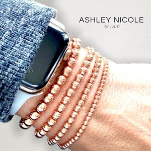 Könnte beinhalten: Ein Set aus fünf roségoldenen Perlenarmbändern, die auf einem Handgelenk übereinander getragen werden. Die Armbänder bestehen aus kleinen, runden Perlen und werden mit einer roségoldenen Apple Watch getragen.