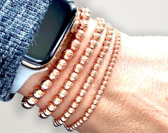 May include: A set of five rose gold beaded bracelets stacked on a wrist. The bracelets are made of small, round beads and are worn with a rose gold Apple Watch.