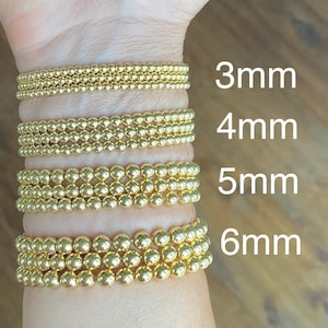 May include: Four gold bracelets with different sized beads. The bracelets are stacked on a person's wrist. The bead sizes are 3mm, 4mm, 5mm, and 6mm.