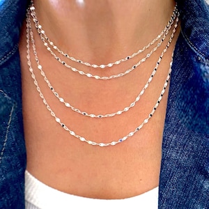 May include: A layered silver necklace with a delicate chain featuring small, oval-shaped links. The necklace is worn by a person, but the focus is on the jewelry itself.