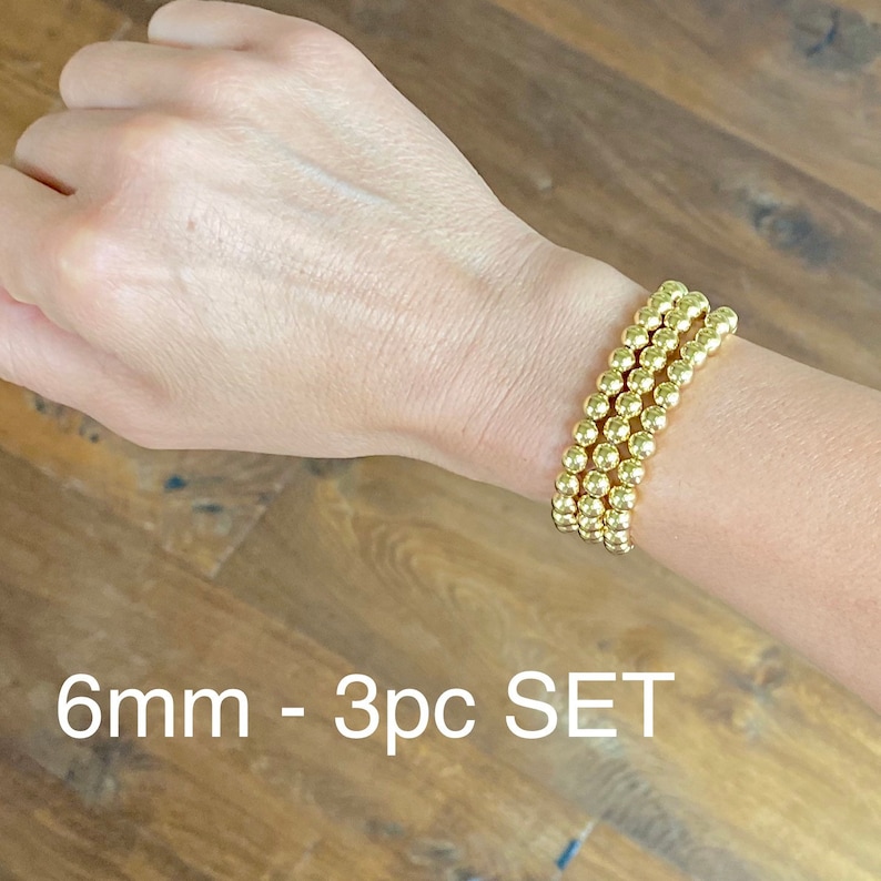 May include: A set of three gold-toned beaded bracelets, each with 6mm beads. The bracelets are stacked on a person's wrist. The text "6mm - 3pc SET" is visible below the bracelets.
