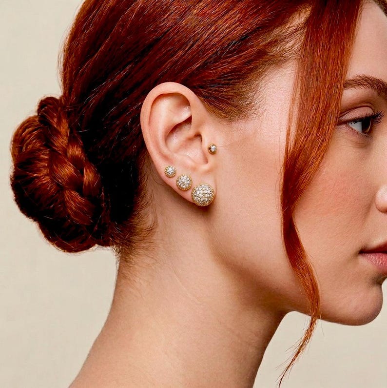 A close-up,-side profile of a female model showcasing three graduated sizes of gold pave disco ball stud earrings. The three round crystal-encrusted studs are worn sequentially along the earlobe in 6mm, 8mm, and 10mm sizes, catching the light with a brilliant, metallic gold finish.