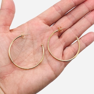 Puede incluir: Un par de pendientes de aro dorados. Los pendientes son circulares con un acabado liso y pulido. Los postes son visibles, lo que indica un diseño para orejas perforadas. Los pendientes se muestran sobre un fondo claro.