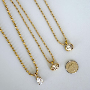 Three 18K PVD gold ball chain CZ pendant necklaces laid on marble surface showing oval marquise and pear cubic zirconia pendant styles side by side