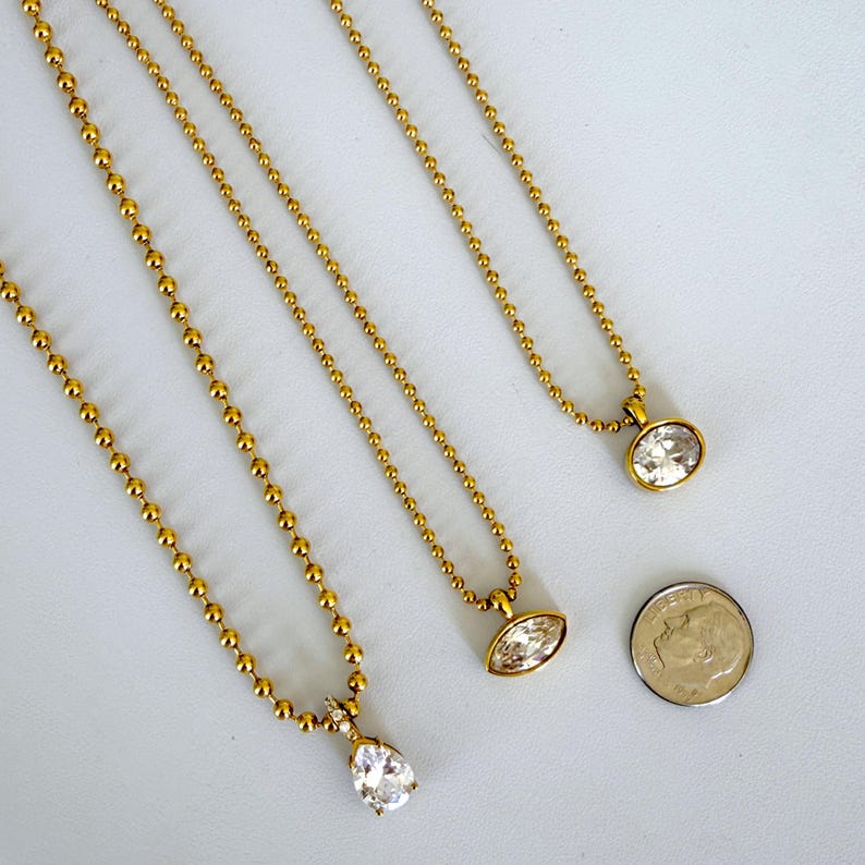 Three 18K PVD gold ball chain CZ pendant necklaces laid on marble surface showing oval marquise and pear cubic zirconia pendant styles side by side