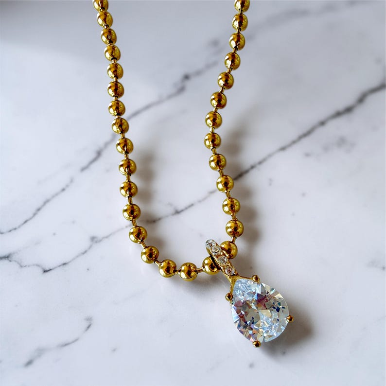 Close up of Pear cubic zirconia teardrop pendant necklace on white marble showing 18K PVD gold 3mm ball chain with pavé bail and sparkling CZ stone