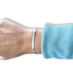 Puede incluir: Una pulsera de plata en la muñeca de una persona. La pulsera es fina y plana, y tiene un acabado pulido.