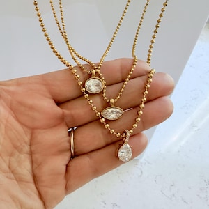 Three 18K PVD gold ball chain necklaces held in palm showing oval, marquise, and pear cubic zirconia pendant styles on warm yellow gold ball chain, shower safe jewelry