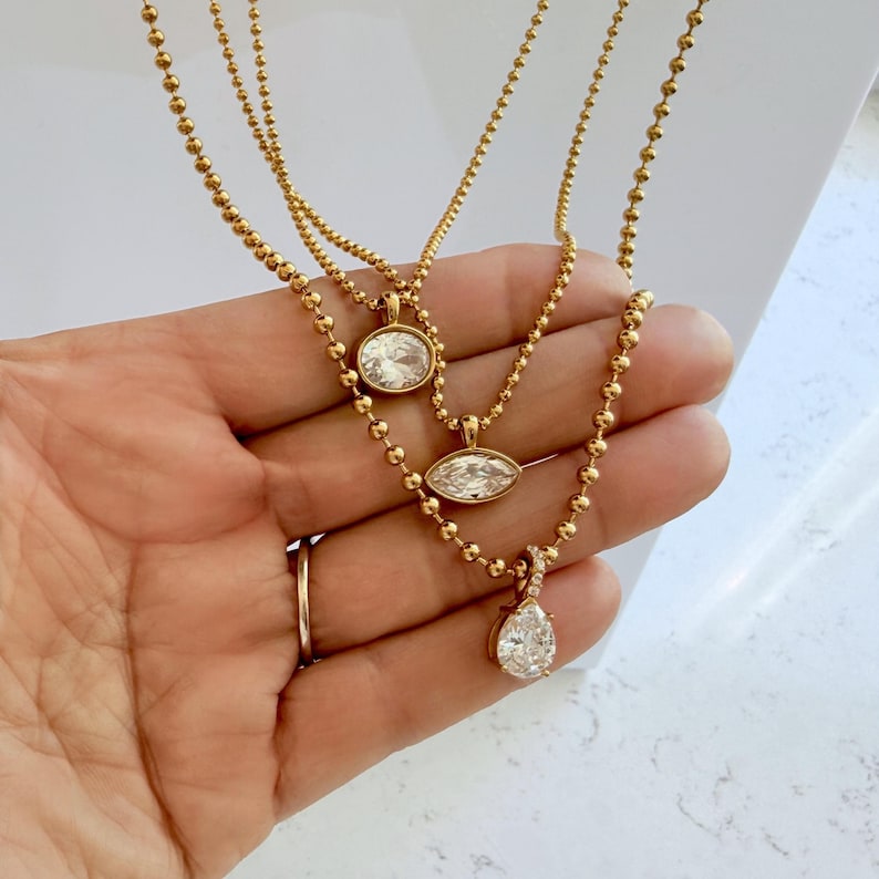 Three 18K PVD gold ball chain necklaces held in palm showing oval, marquise, and pear cubic zirconia pendant styles on warm yellow gold ball chain, shower safe jewelry